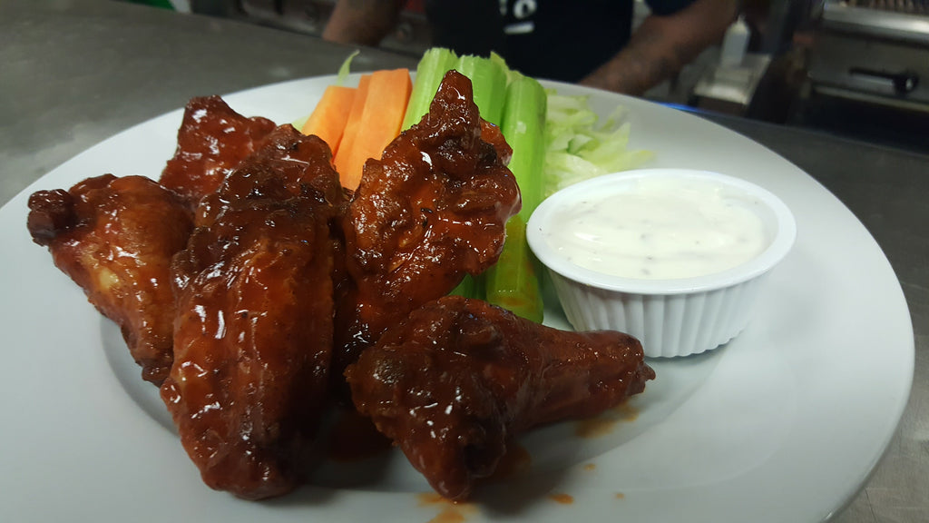 A plate of buffalo wing wings in a glossy reddish-brown sauce, served with celery sticks, carrot sticks, and a small ramekin of white dipping sauce on a white plate.