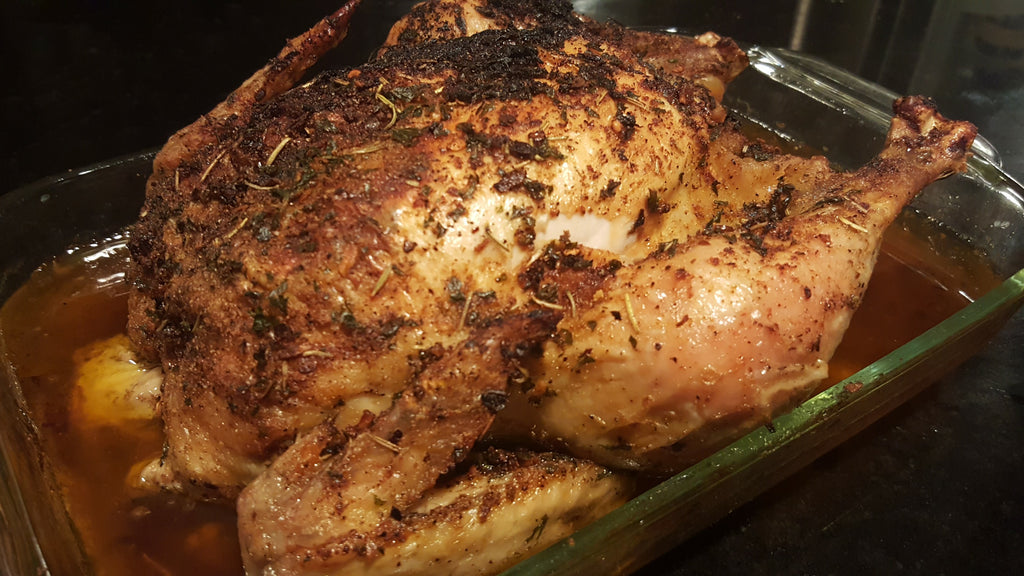 A whole roasted chicken with a golden-brown crispy skin and herb seasoning, sitting in a glass baking dish with a browned liquid base.